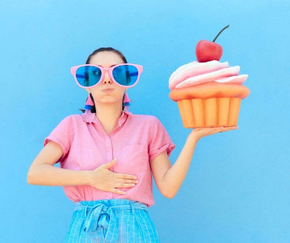 woman wearing sunglasses holding her stomach with one hand and a large cupcake with the other